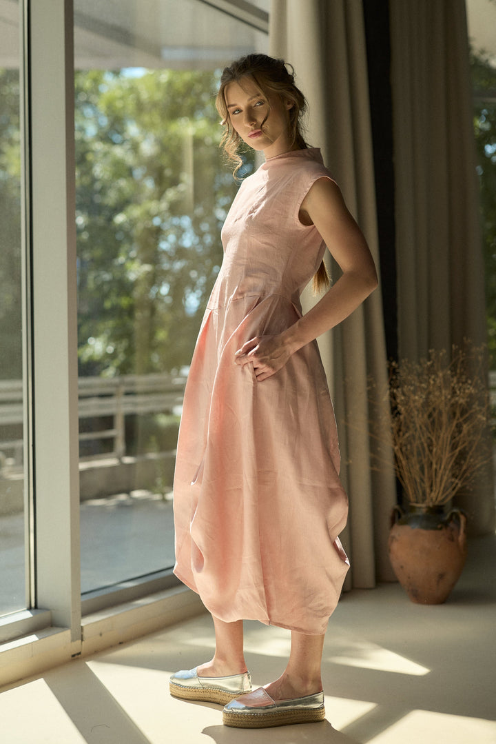 A woman wearing a sleeveless, high-neck, midi-length dusty pink dress with a gathered, draped silhouette, standing in a brightly lit room next to a large window.