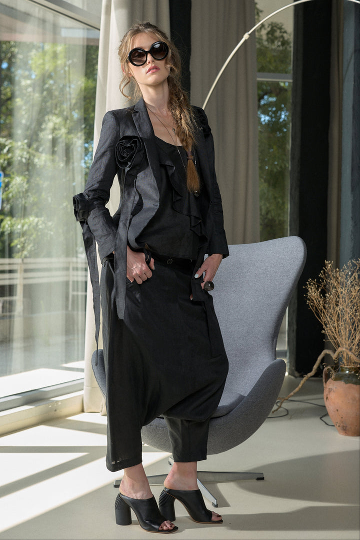 A woman models a black fashion outfit consisting of a structured blazer with rose details, a black top, loose black trousers, and black high-heeled mules, posed in a modern room with a gray chair and large windows.