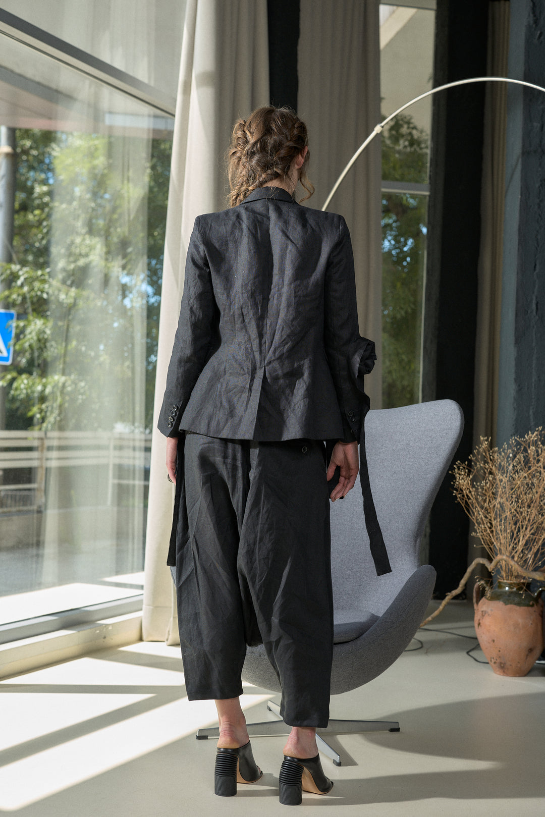A model viewed from behind wearing a dark charcoal gray two-piece suit consisting of a textured blazer and cropped trousers, standing in a modern room next to a gray lounge chair.