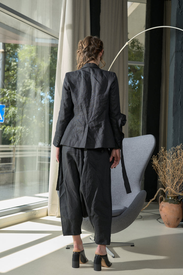 A model viewed from behind wearing a dark charcoal gray two-piece suit consisting of a textured blazer and cropped trousers, standing in a modern room next to a gray lounge chair.