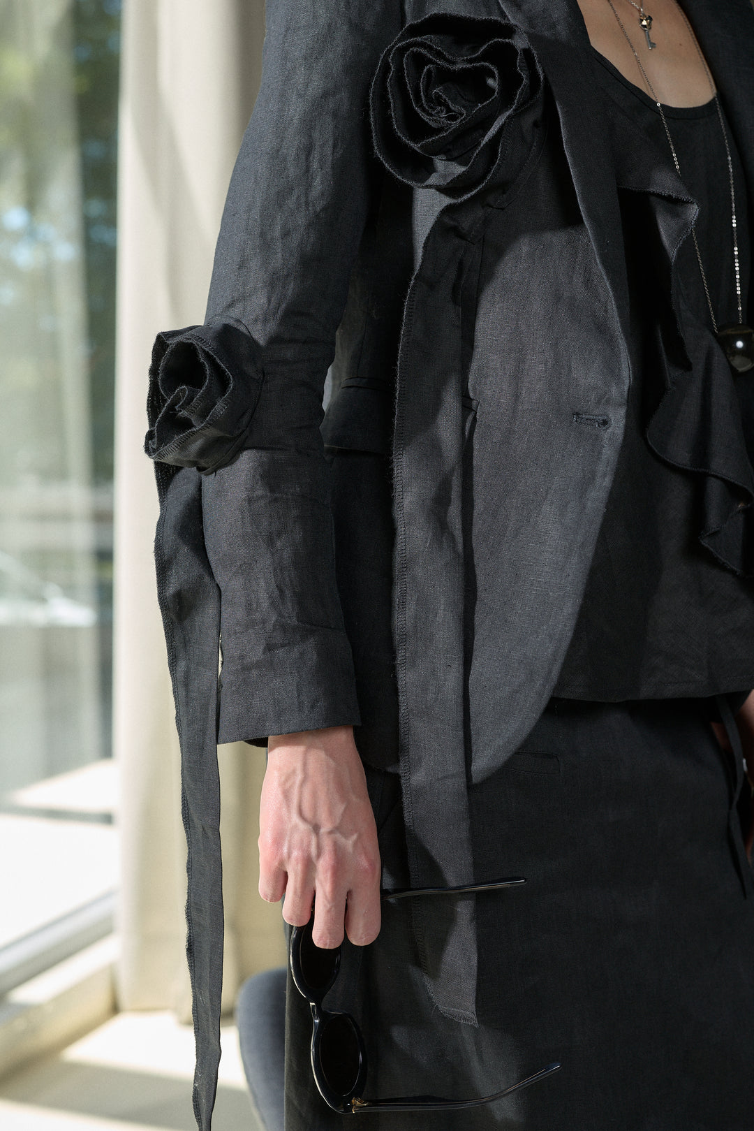 A close-up of a person wearing a dark charcoal gray linen jacket with large sculptural fabric roses on the shoulders and sleeves, paired with a black ruffled top and skirt. The person is holding black sunglasses in front of a bright window.