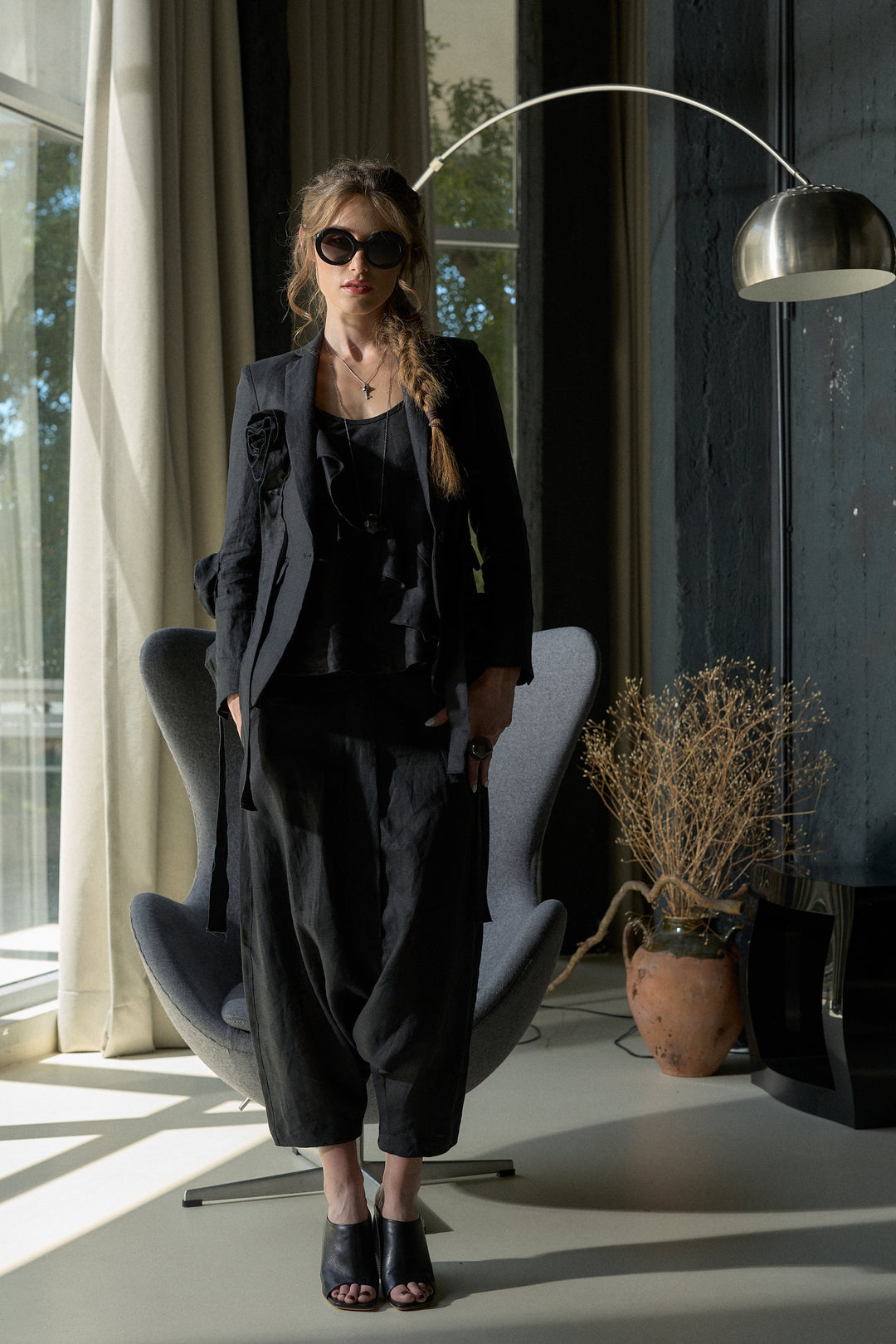 A woman wearing a black ruffled blouse, matching black wide-leg trousers, and black open-toe heels stands in a modern room with a gray lounge chair, a floor lamp, and large windows.