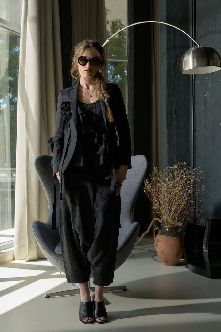 A woman wearing a black ruffled blouse, matching black wide-leg trousers, and black open-toe heels stands in a modern room with a gray lounge chair, a floor lamp, and large windows.