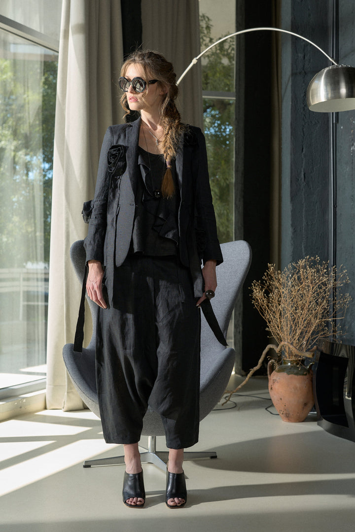 A woman stands in a modern room wearing a black tailored outfit consisting of a blazer, top, and wide-leg cropped trousers, paired with black peep-toe heels. She is positioned in front of a gray chair, with a silver floor lamp and a potted dried plant in the background.