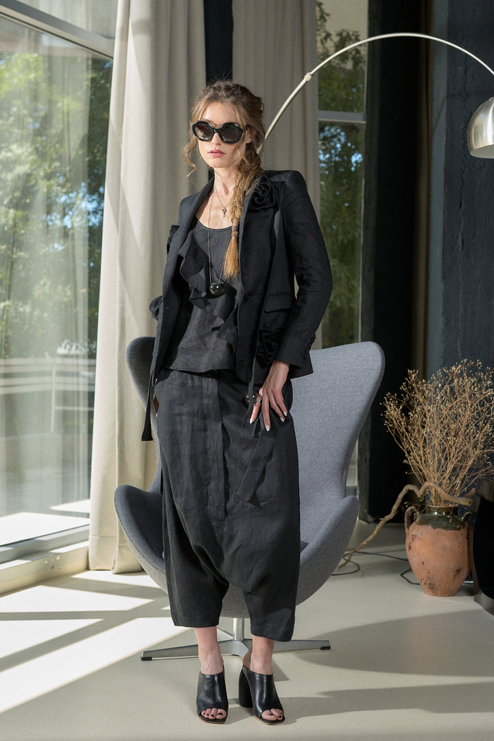A woman models a black two-piece suit consisting of a cropped blazer and matching pleated trousers, styled with black sunglasses, a black top, and black high-heeled sandals in a modern room setting.