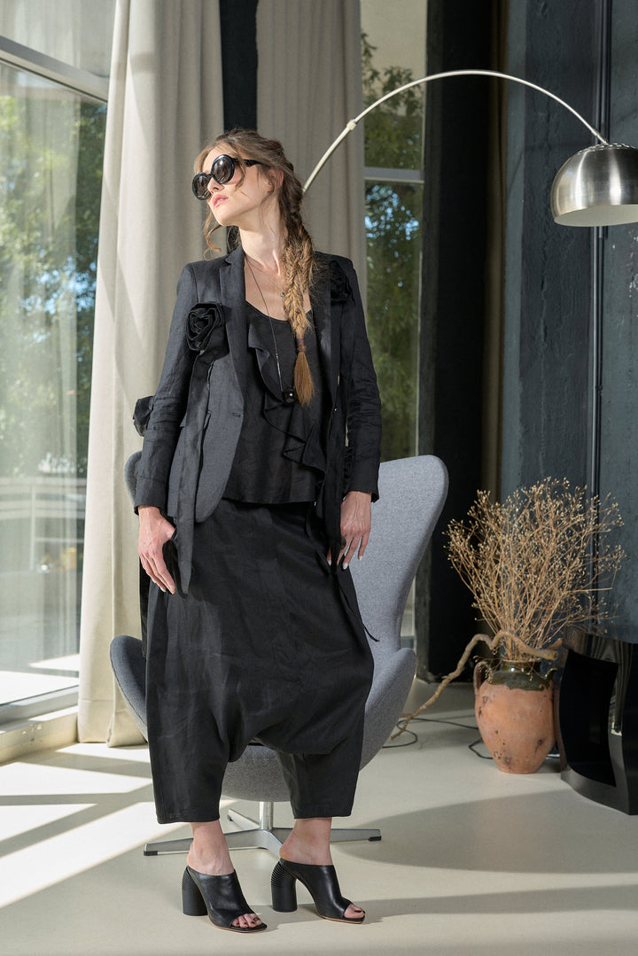 A woman models a black ruffled longline vest and matching wide-leg trousers with black open-toe heels in a modern room setting with a gray chair and a silver floor lamp.