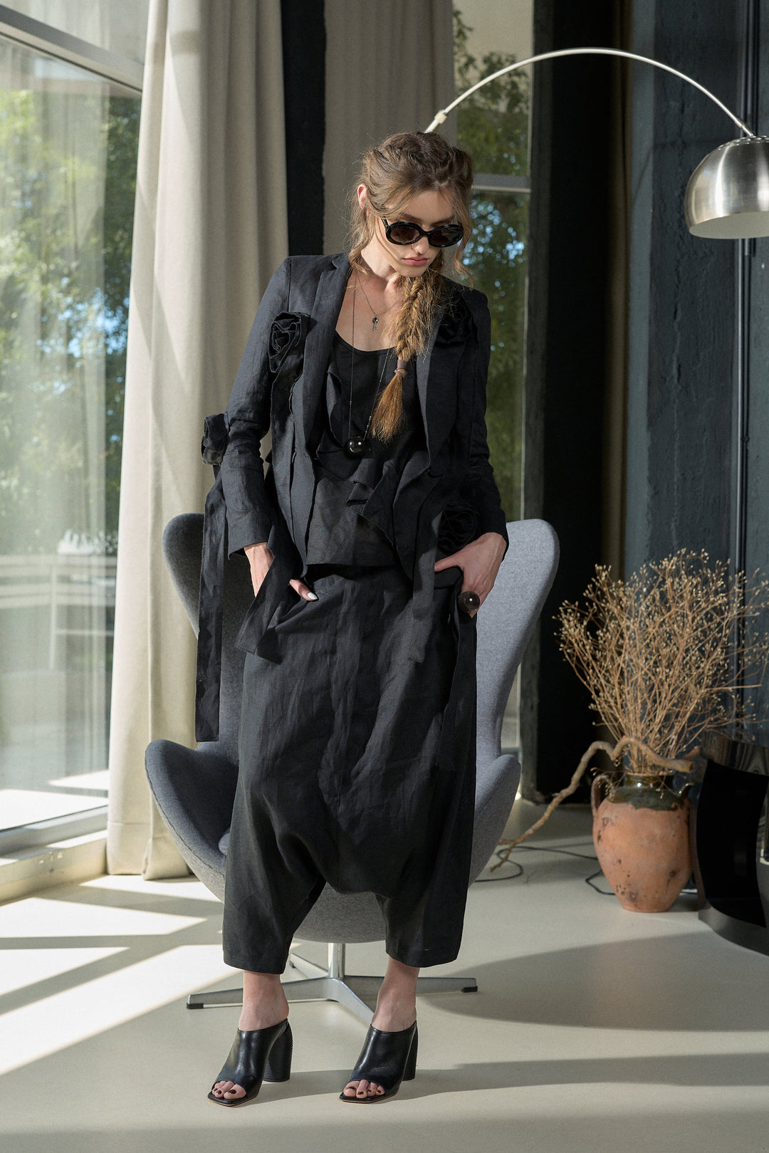 A woman models a black long-sleeve linen-blend jumpsuit with a relaxed, baggy fit and ruffled details on the shoulders, styled with black sunglasses, a black top, and black peep-toe heels. She is seated in a modern gray lounge chair inside a stylishly decorated room with large windows, a floor lamp, and a potted plant.
