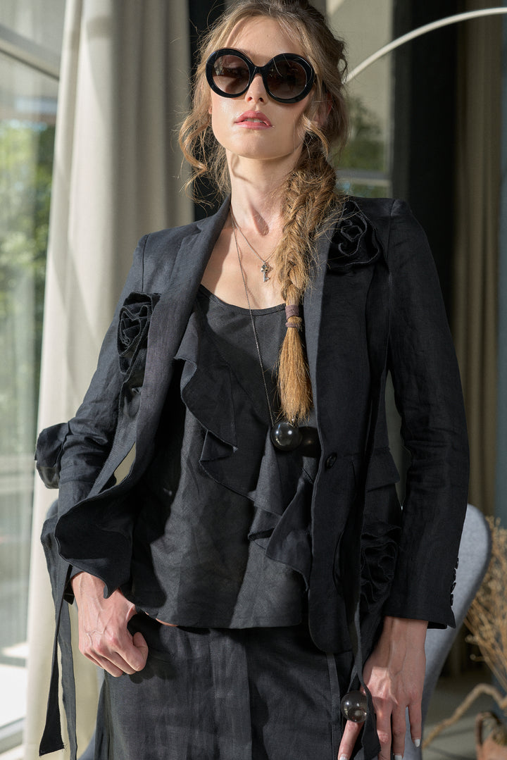 A model wearing a dark gray ruffled blouse, a matching dark gray ruffled blazer with decorative fabric flowers on the shoulders, matching trousers, and oversized black sunglasses.