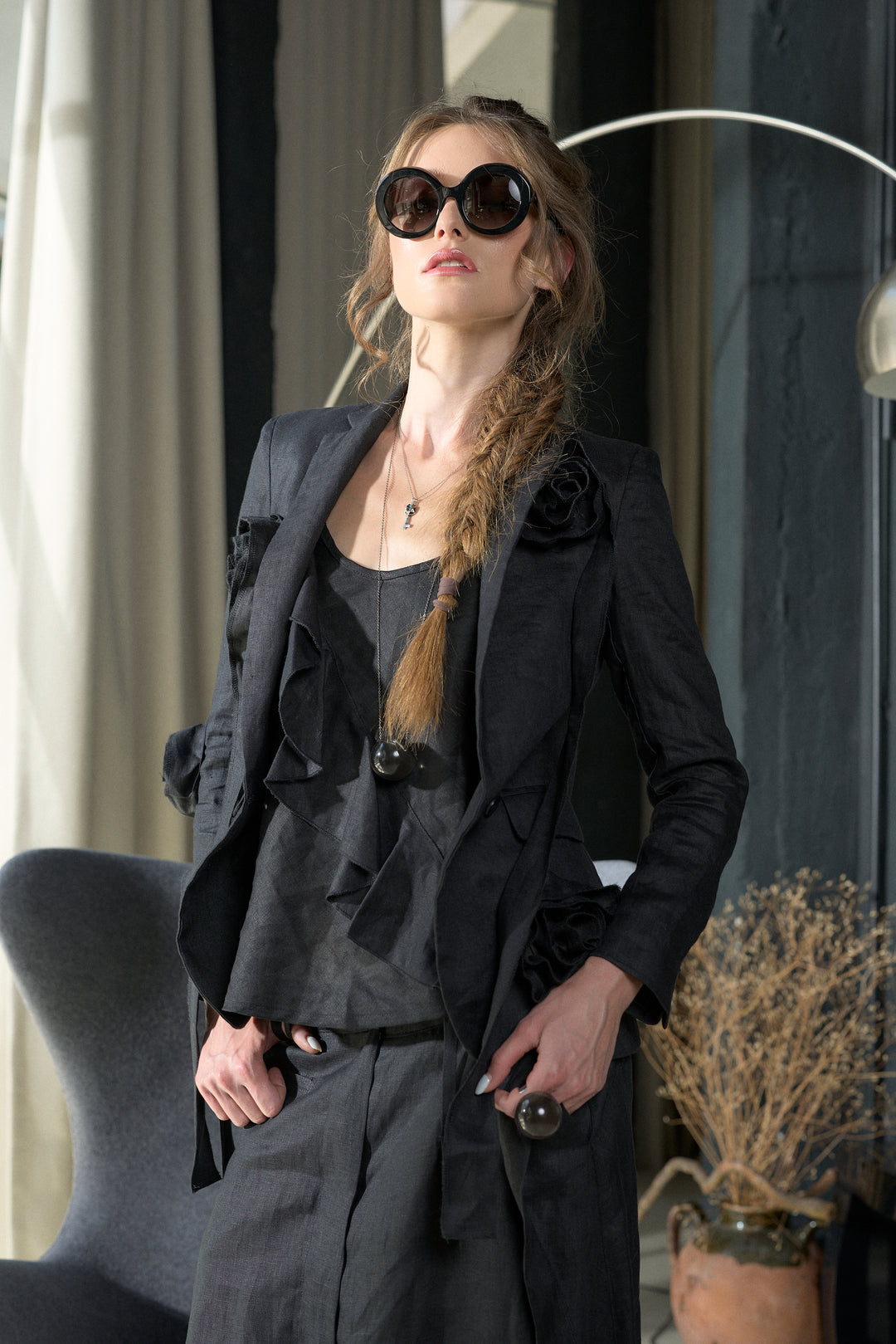 A woman wearing a dark ruffled jacket, matching top and trousers, oversized dark sunglasses, and a necklace, standing indoors near a chair and potted dried plants.
