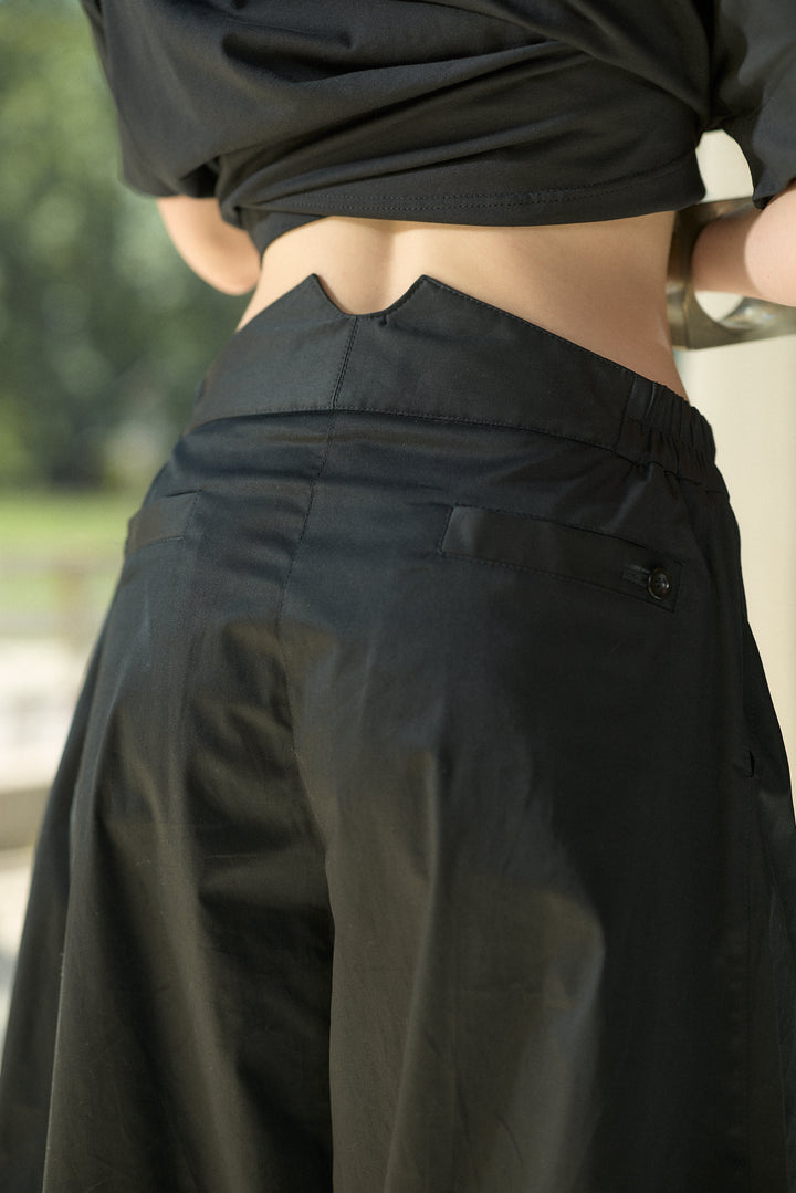 Close-up back view of a person wearing black wide-leg pants and a black cropped top, with a blurred outdoor background.