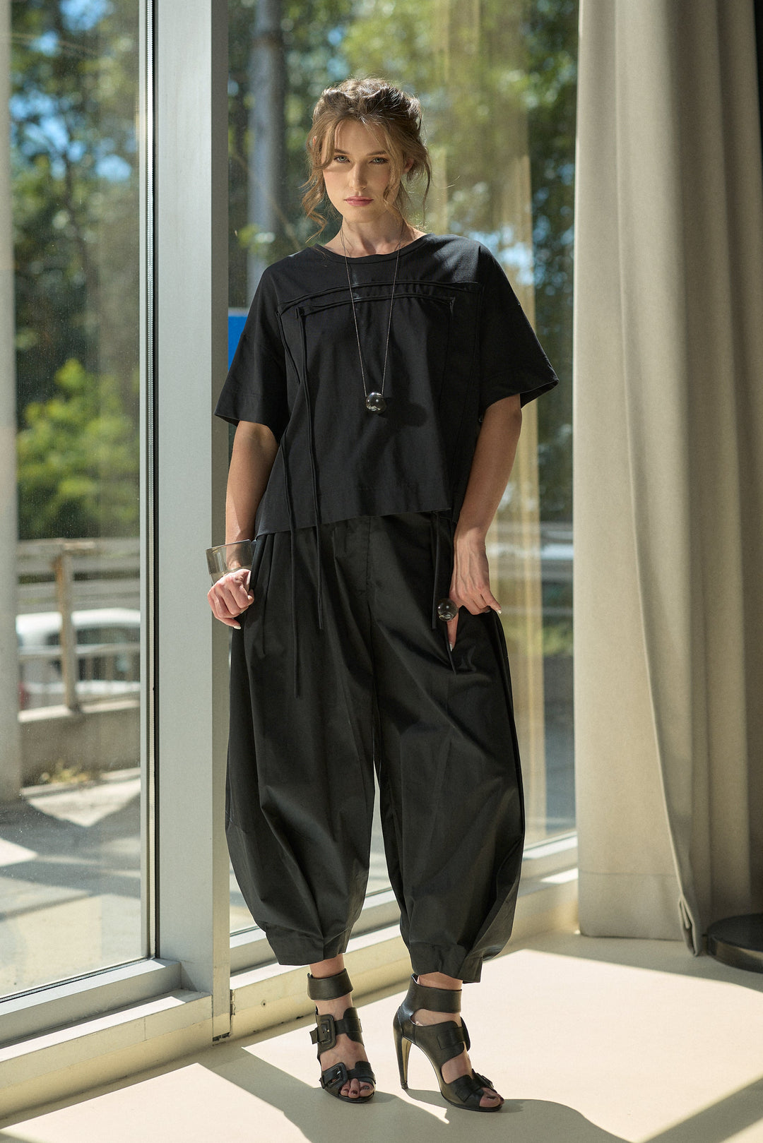 A woman stands indoors by large glass windows wearing a black short-sleeve top with a gathered neckline and matching loose black wide-leg pants, paired with black strappy heels.