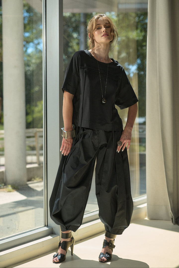 A woman models a black two-piece outfit consisting of a short-sleeve top and matching wide-leg harem pants, standing in front of a large window with natural light.