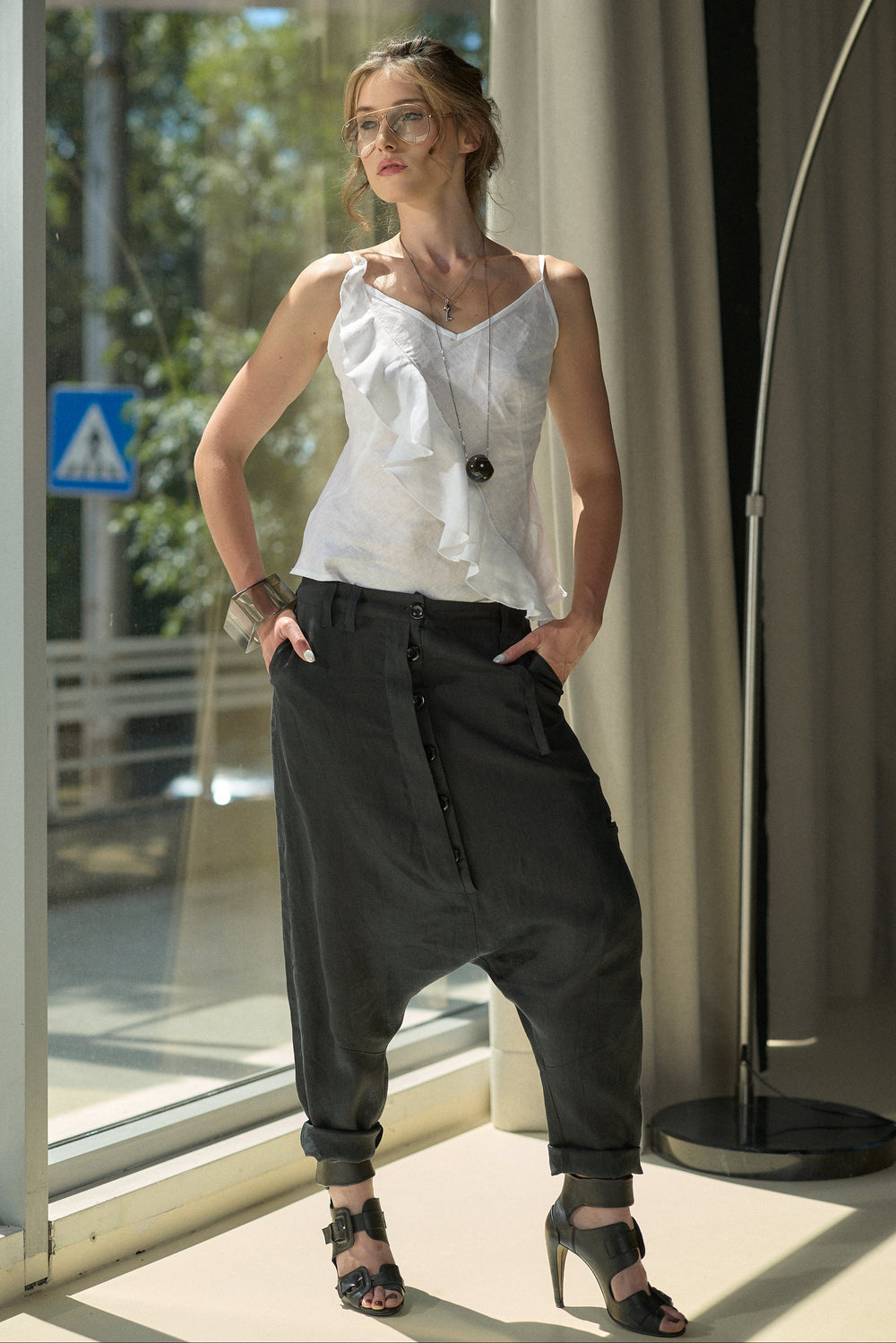 A woman modeling a white ruffled sleeveless top and black baggy cuffed pants with black high-heeled sandals, standing in front of a large window with curtains and a floor lamp in a bright indoor setting.