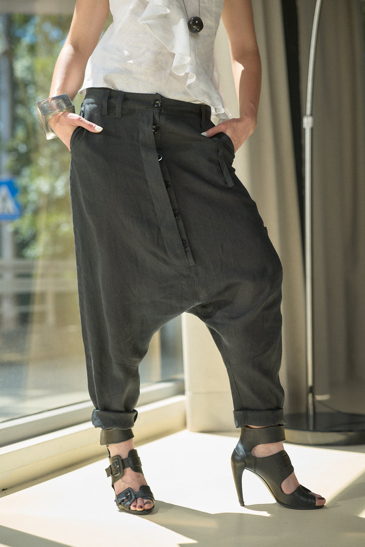 A person wearing loose black drape pants with rolled cuffs, paired with a white ruffled top and black high-heeled sandals, standing near a window.