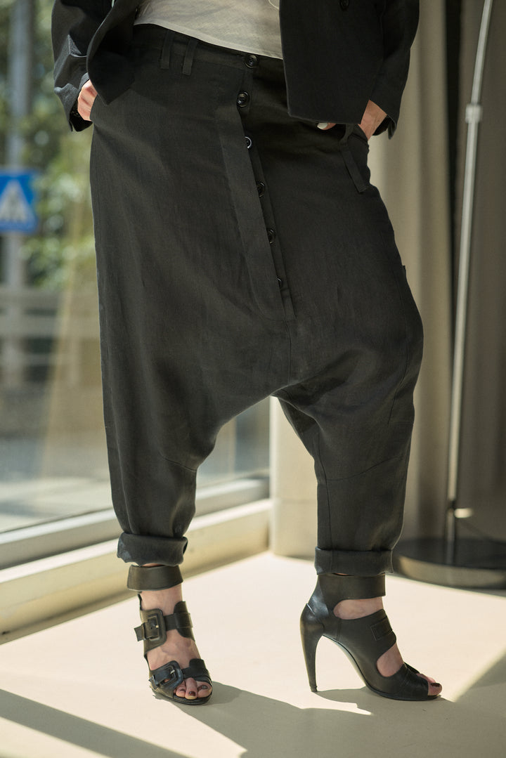 A person wearing loose black drape pants with rolled cuffs, paired with black high-heeled sandals and a black blazer, standing indoors near a window.