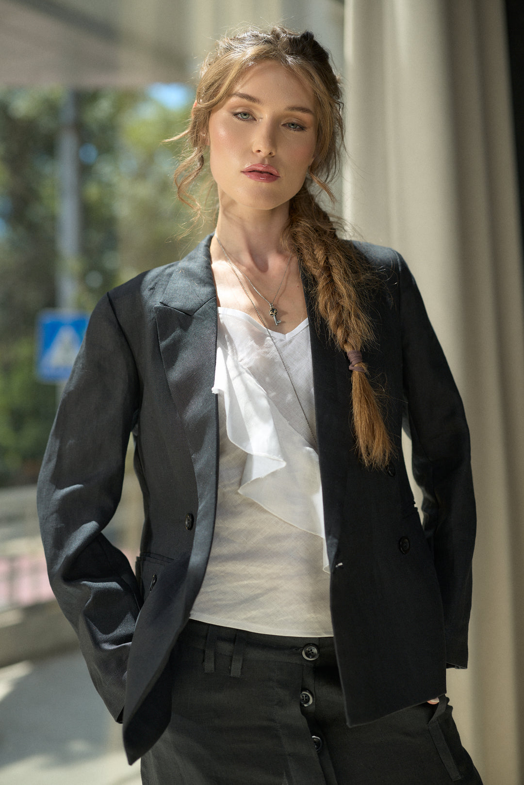 A woman wearing a black double-breasted blazer over a white ruffled top and black trousers, standing in a bright, modern indoor-outdoor space.