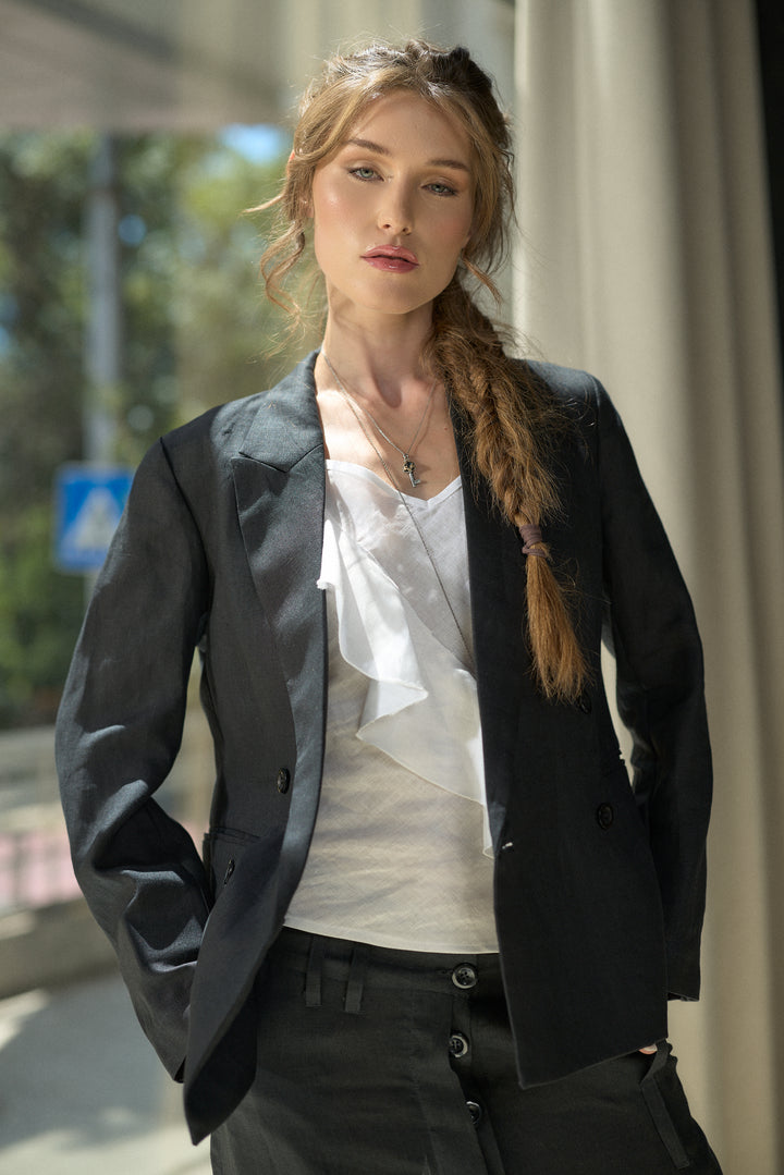 A woman wearing a black double-breasted blazer over a white ruffled top and black trousers, standing in a bright, modern indoor-outdoor space.