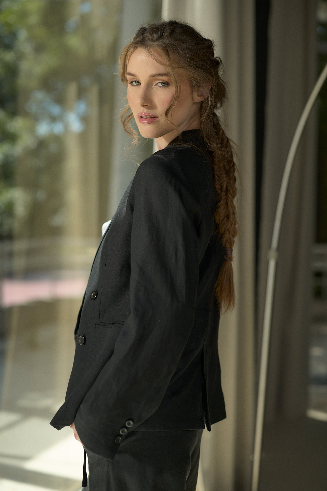 A woman with a long brown braid wearing a black double-breasted blazer and matching trousers, posing in profile against a softly lit modern interior background.