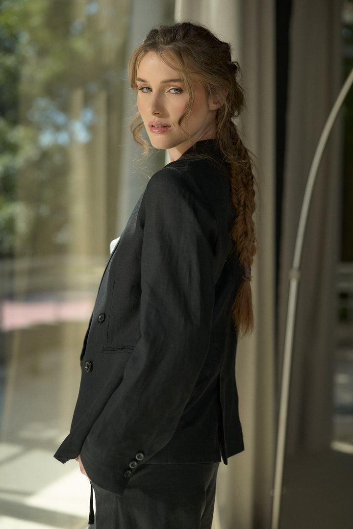 A woman with a long brown braid wearing a black double-breasted blazer and matching trousers, posing in profile against a softly lit modern interior background.
