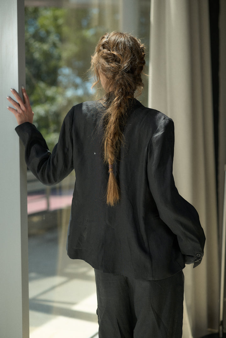 A person viewed from behind wearing a black oversized blazer and matching pants, standing indoors near a window and touching the frame. The hair is styled in a long braided ponytail.