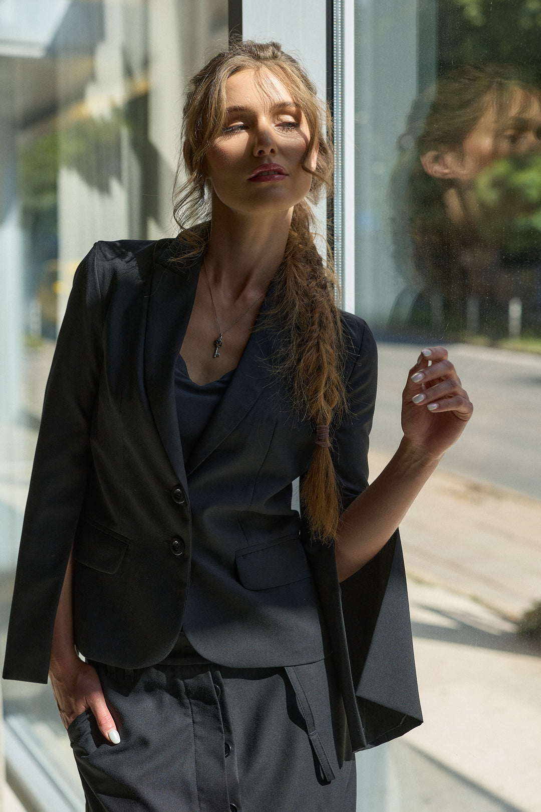A woman with a long blonde braid stands by a window wearing a dark charcoal gray tailored blazer and matching trousers over a dark top.