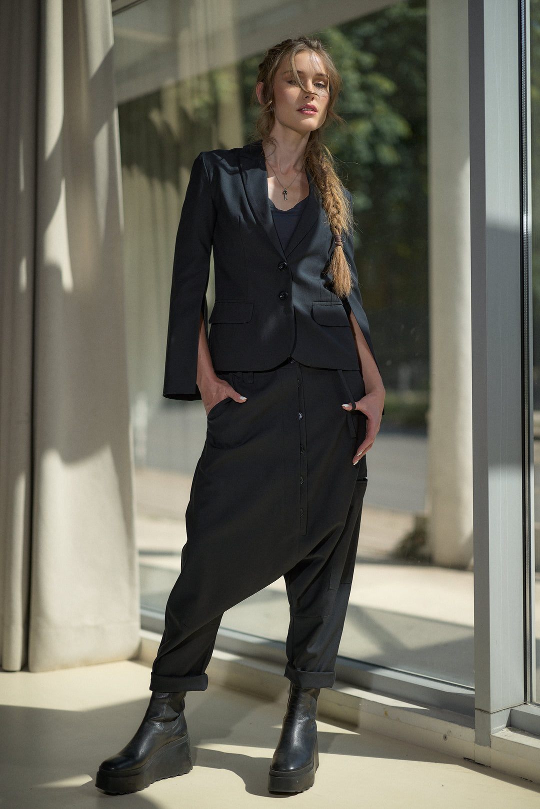 A woman models a dark navy two-piece suit consisting of a tailored blazer and matching relaxed trousers, paired with black platform boots, standing in front of a modern glass window.
