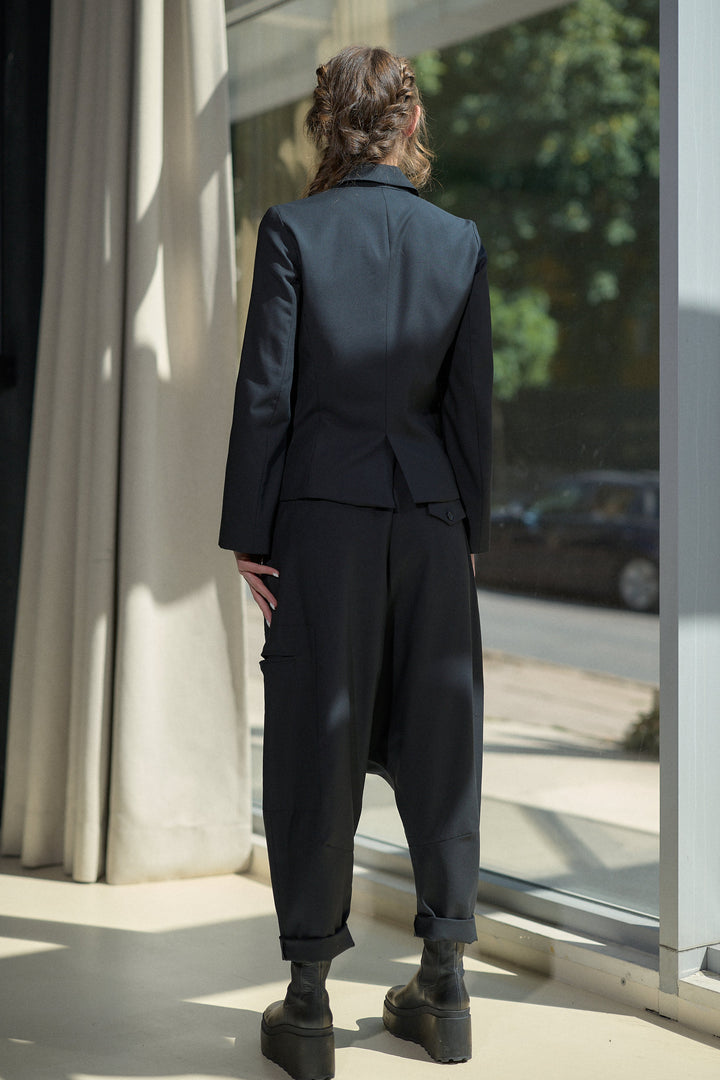 A person viewed from behind wearing a dark tailored suit with a structured blazer and loose cropped trousers, standing indoors near large windows and curtains.