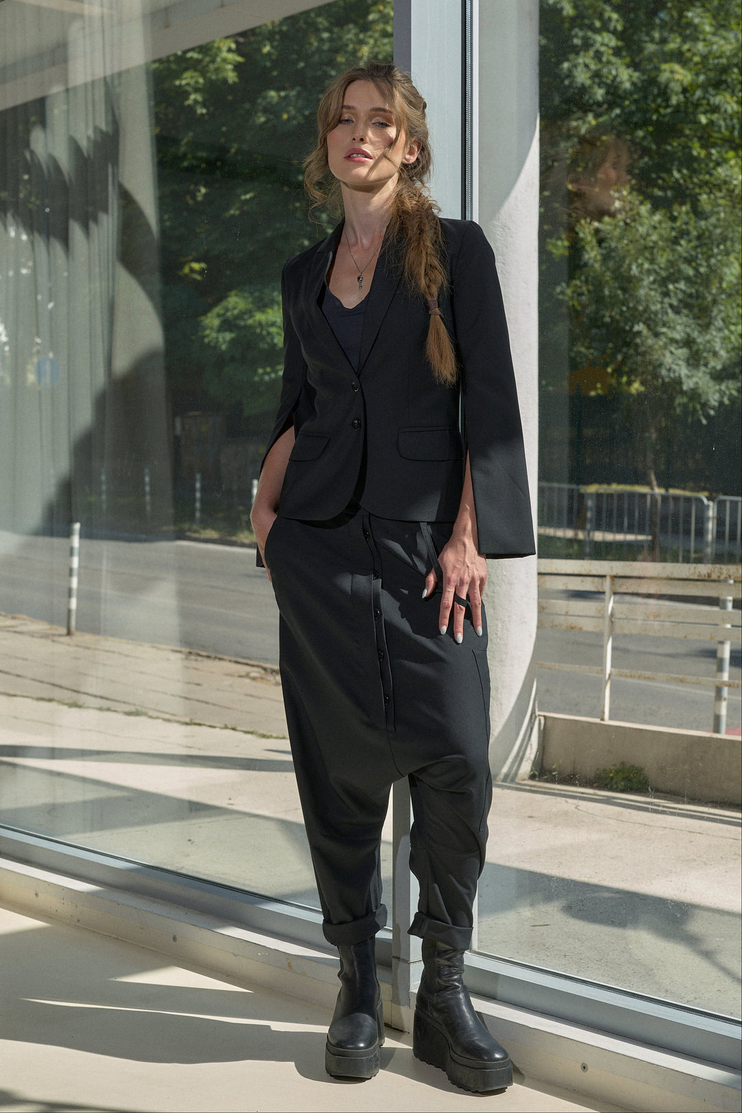 A woman stands outdoors beside a glass building, wearing a black tailored blazer with three-quarter sleeves, matching loose black trousers, and black ankle boots.