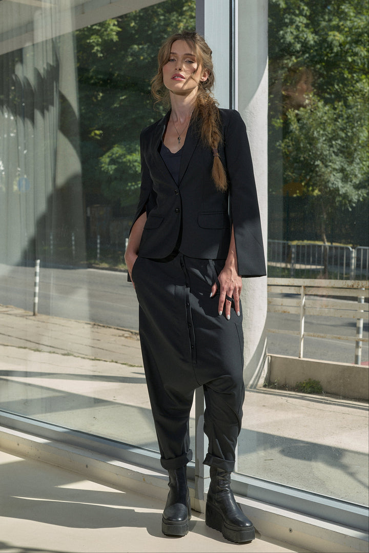 A woman stands outdoors beside a glass building, wearing a black tailored blazer with three-quarter sleeves, matching loose black trousers, and black ankle boots.