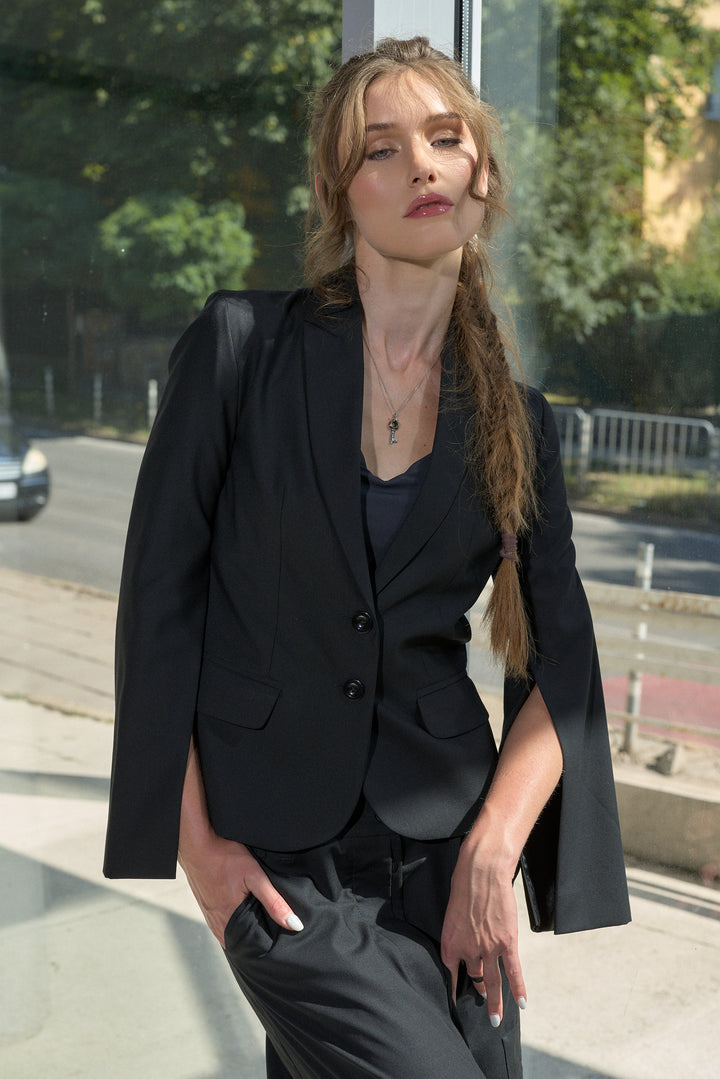 A woman wearing a black structured blazer with dramatic shoulder flaps and black trousers, standing outdoors in front of a glass building.