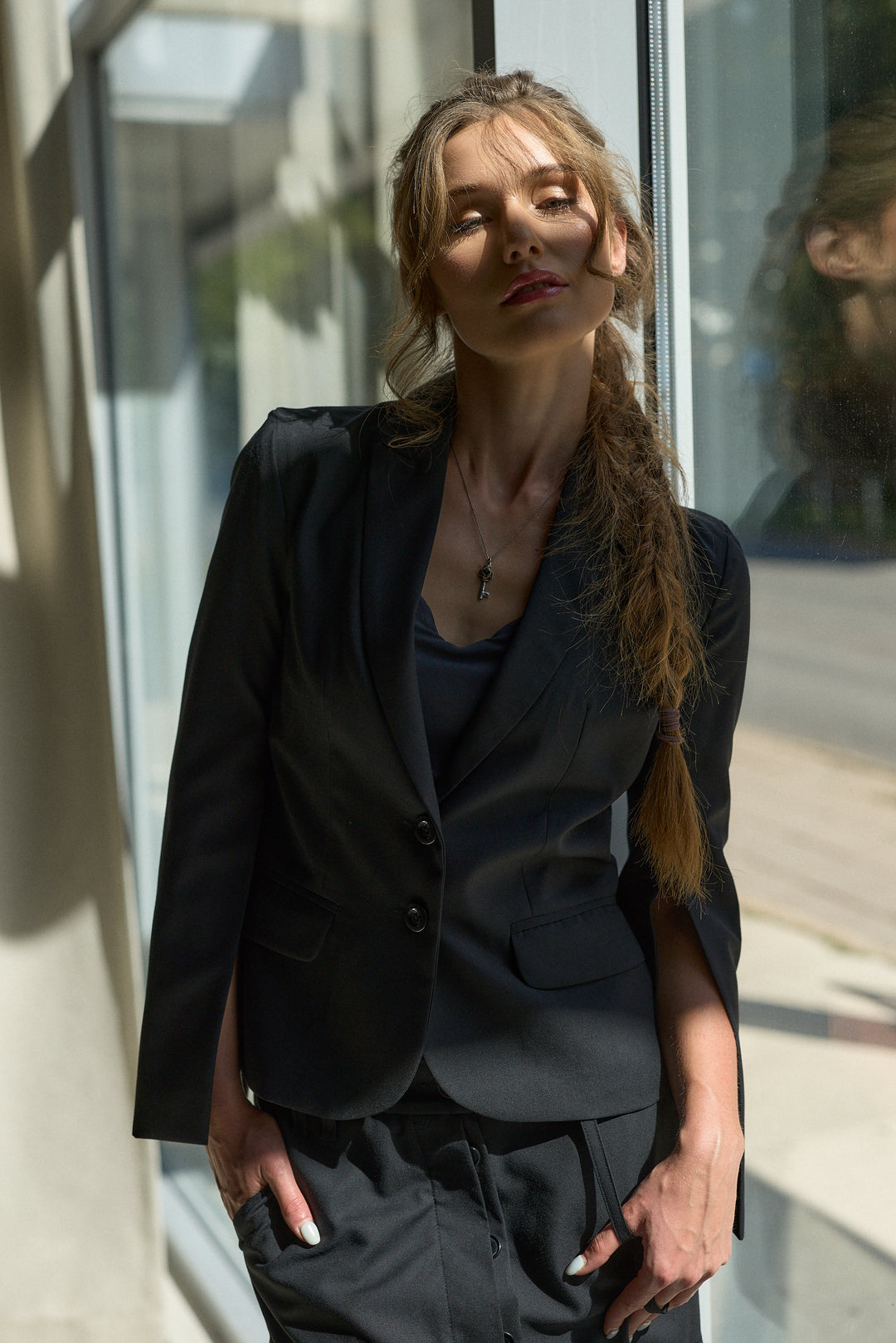 A woman wearing a black tailored blazer and matching dark trousers, standing in front of a window with natural light casting shadows on the wall.