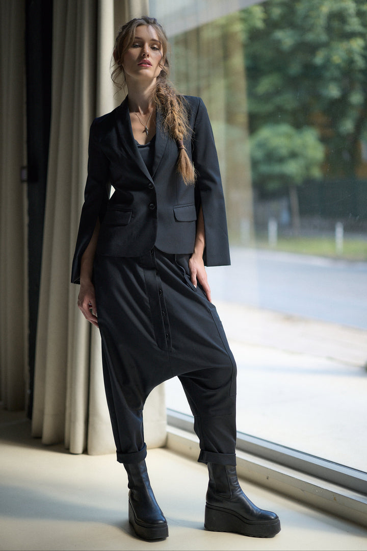 A woman models a black two-piece suit consisting of a cropped blazer with wide sleeves and matching relaxed-fit pants with a drawstring, paired with black platform ankle boots, standing by a large window.