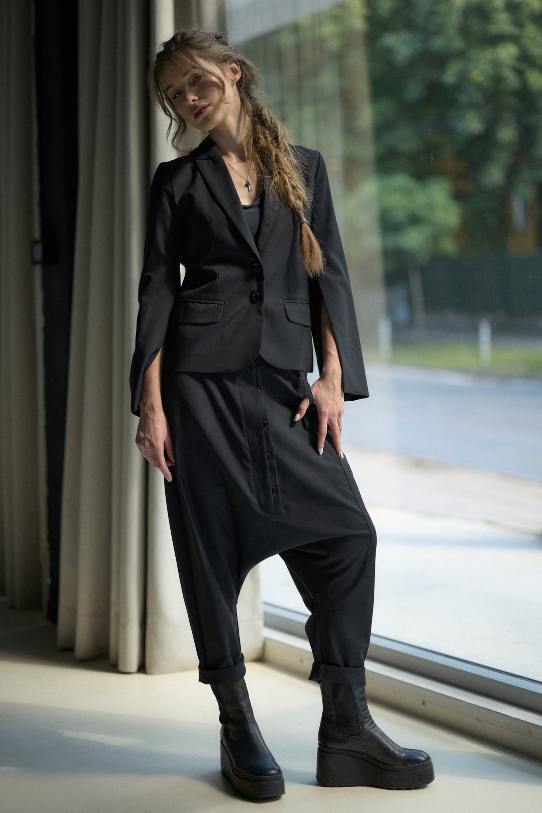 A woman models a black two-piece outfit consisting of a cropped blazer with wide cape-like sleeves and matching relaxed cargo-style pants, paired with black platform ankle boots, standing by a large window.