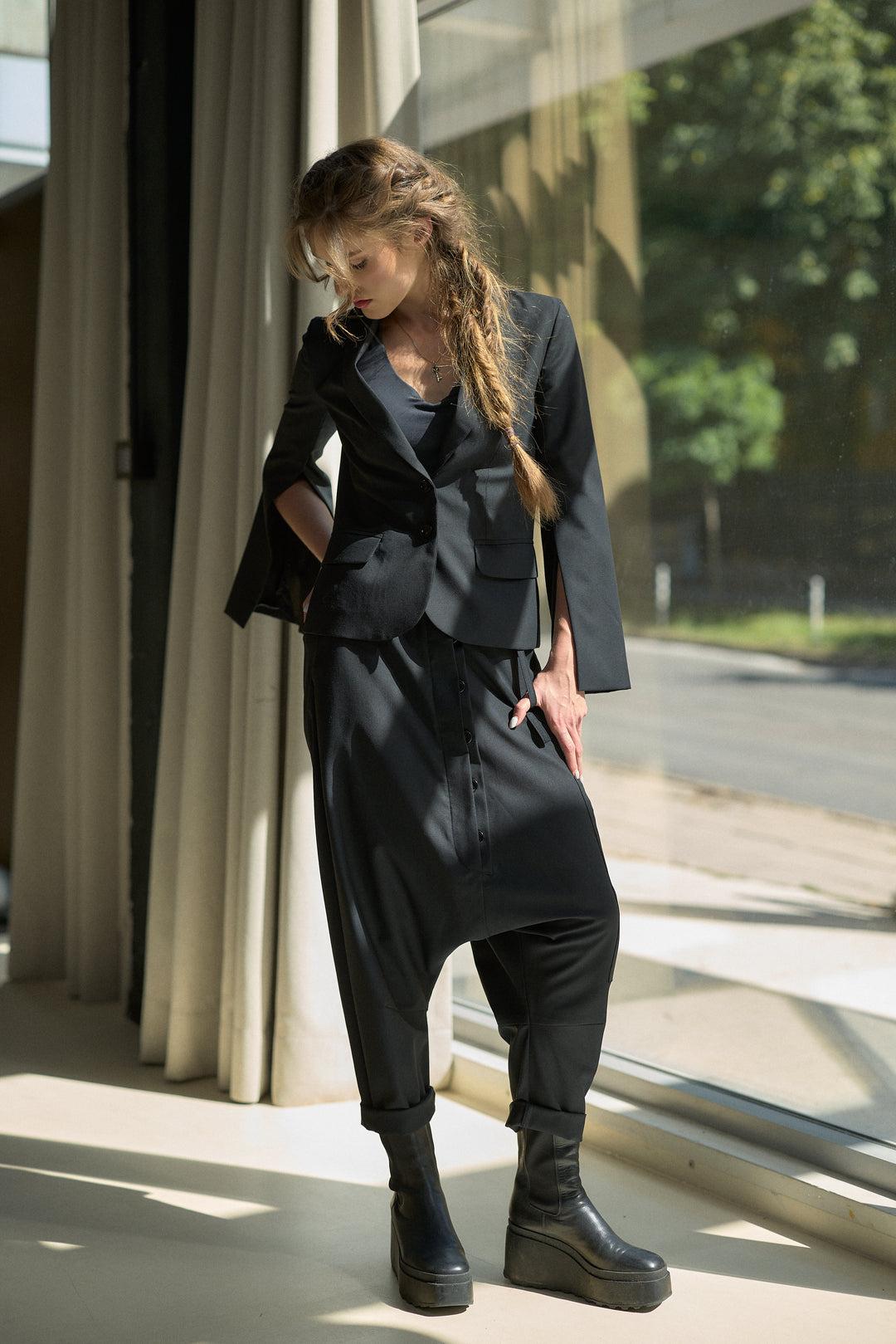 A woman stands by a large window wearing a black tailored outfit with a structured blazer, loose draped pants, and black platform boots.