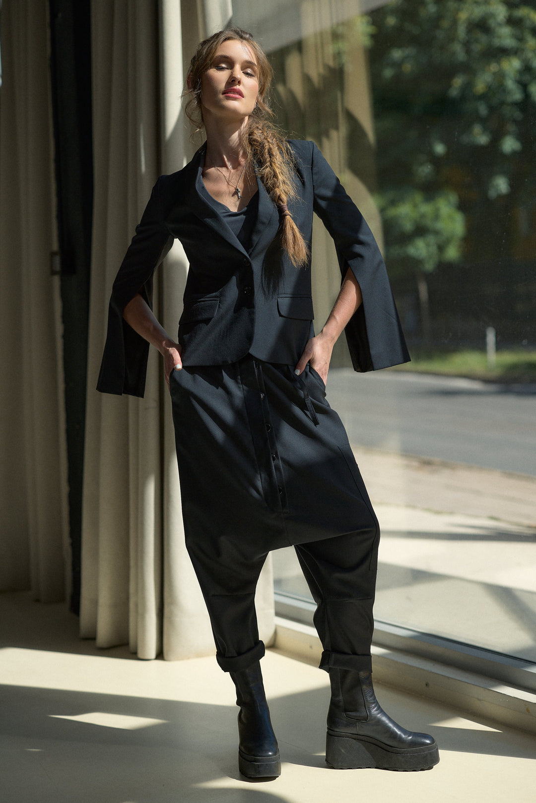 A woman models a black two-piece outfit consisting of a cropped blazer with dramatic wide sleeves and matching loose cuffed pants, paired with black platform boots, standing in front of a window with curtains.