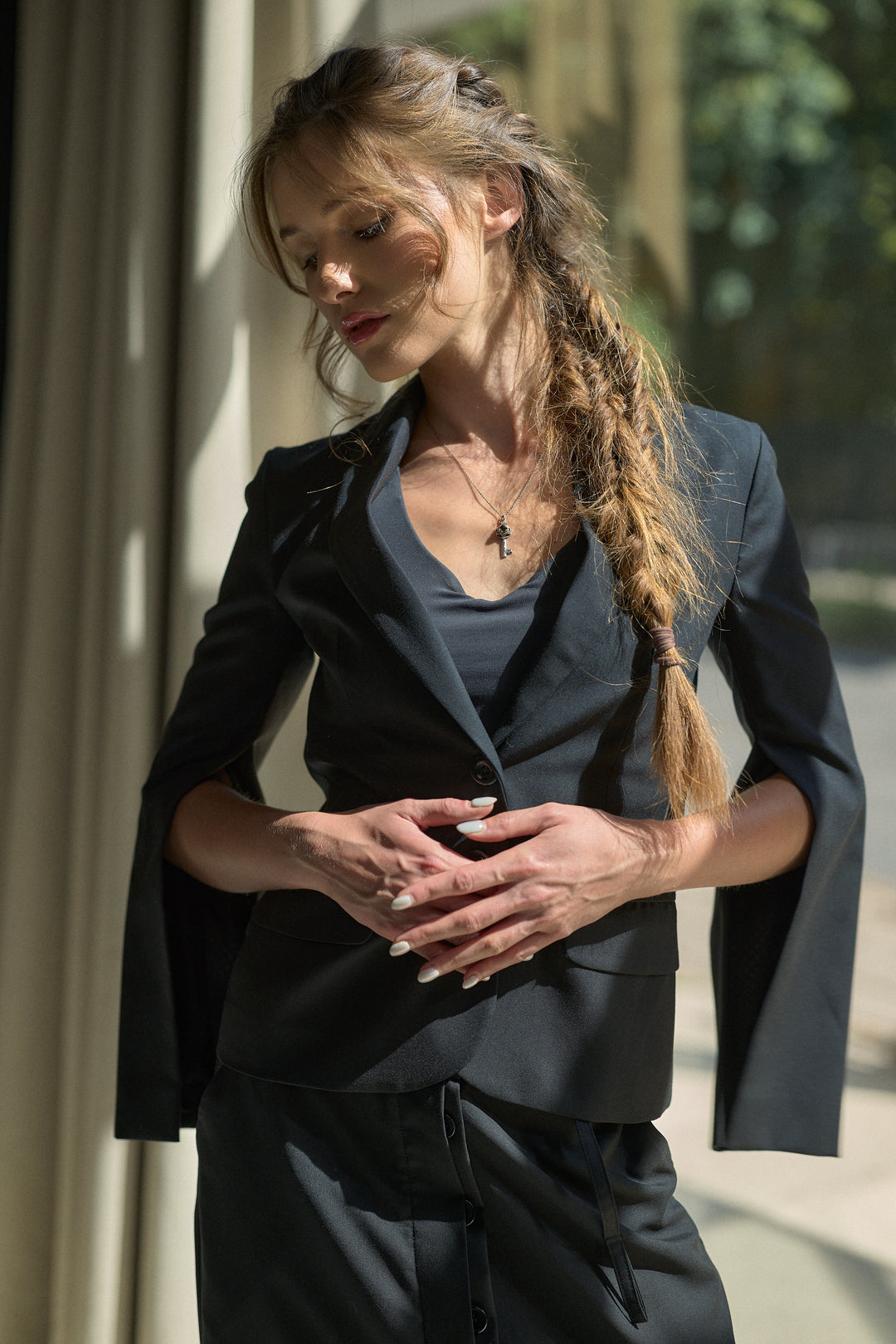 A woman wearing a dark charcoal gray tailored blazer and matching drawstring trousers, styled with a dark gray top and a cross necklace, standing in a sunlit outdoor corridor.