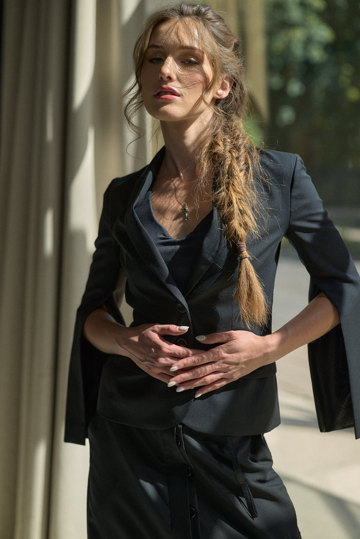 A woman wearing a black tailored blazer with wide lapels and three-quarter sleeves, paired with a matching black top and skirt, standing in a sunlit architectural setting.