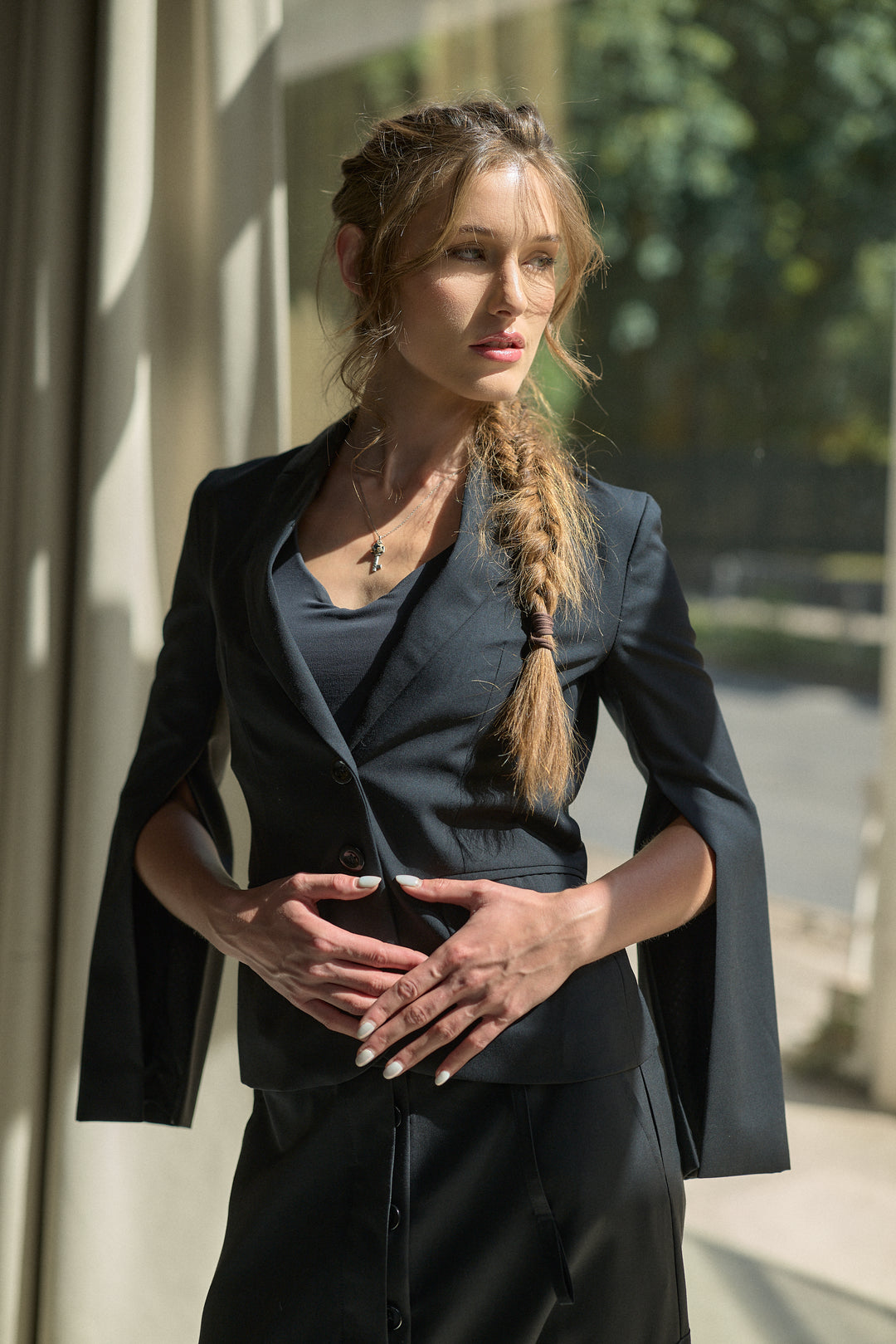 A woman with a long blonde braid stands outdoors near a building, wearing a dark tailored blazer with wide cape-like sleeves over a dark top and skirt. The photo is shot in warm natural light with a softly blurred background of greenery and architecture.