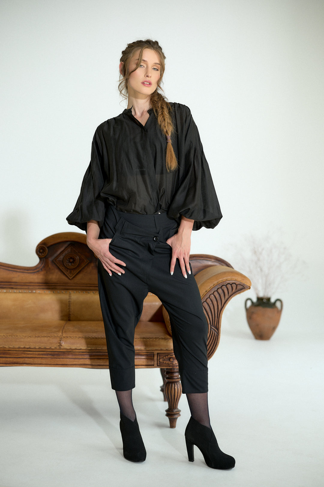 A woman models a black satin blouse with dramatic puffed sleeves and black cropped trousers, posing on a vintage wooden bench against a plain white studio background.
