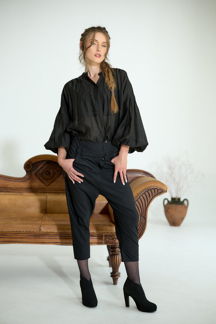 A woman models a black satin blouse with dramatic puffed sleeves and black cropped trousers, posing on a vintage wooden bench against a plain white studio background.
