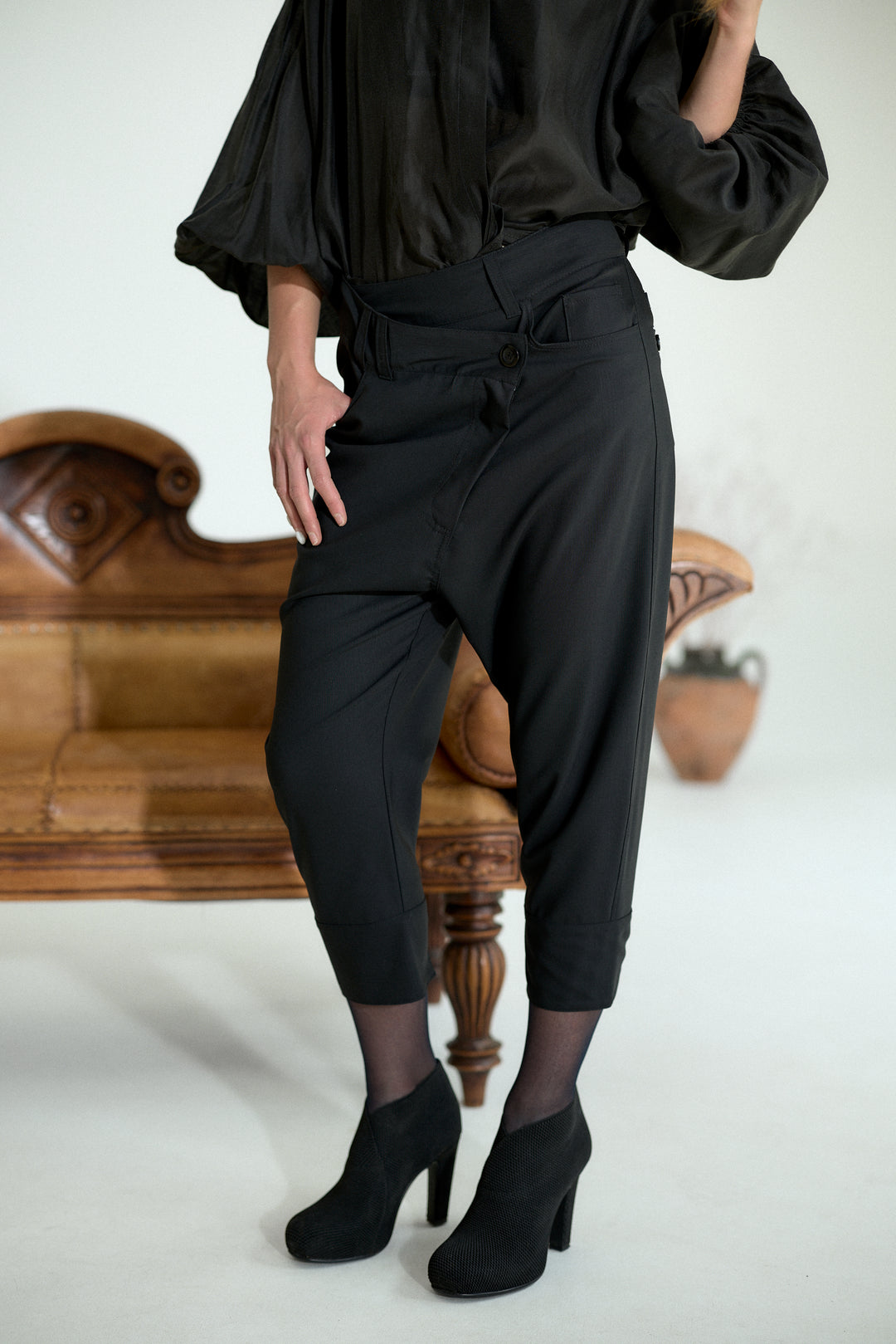 A model wearing black high-waisted cropped trousers with a tie waist, styled with a black blouse and black ankle boots, standing in front of a vintage wooden bench.
