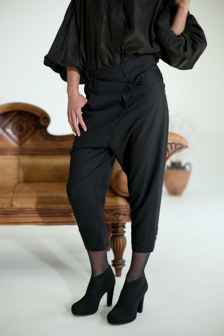 A model wearing black high-waisted cropped trousers with a tie waist, styled with a black blouse and black ankle boots, standing in front of a vintage wooden bench.