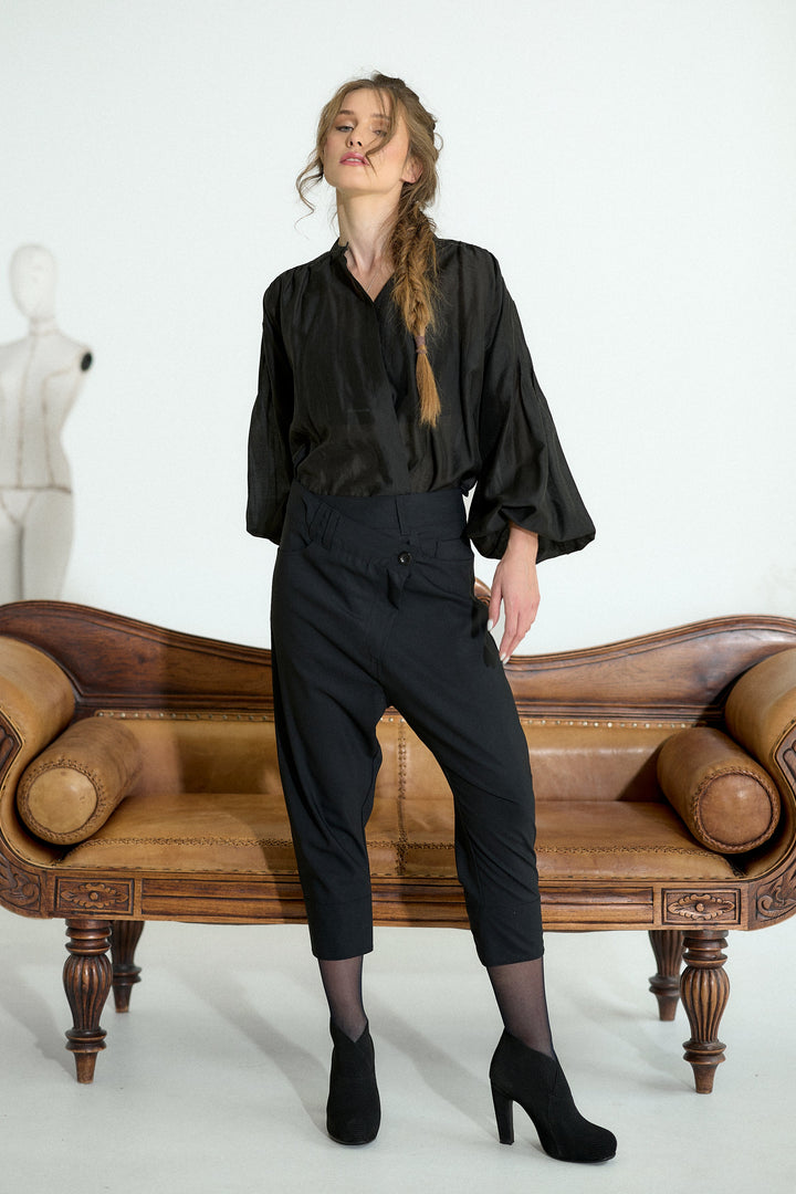 A woman models a black satin blouse and black cropped trousers with sheer black tights and black heels, standing in front of a brown leather bench against a white studio background.