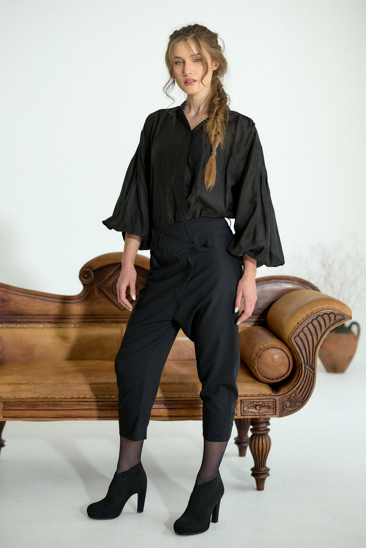 A woman models a black satin blouse with dramatic ruffled sleeves and black cropped trousers, standing in front of a vintage wooden chaise lounge against a white studio background.