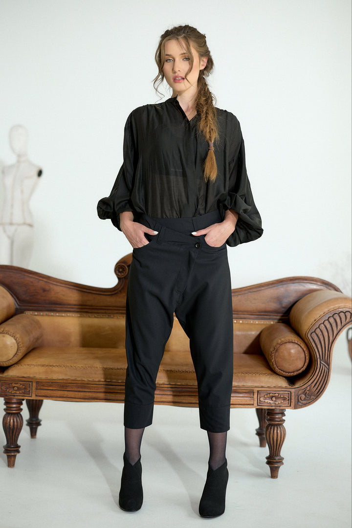 A woman stands in a studio wearing a black long-sleeve blouse and black cropped pants, posed in front of a brown leather bench. The background is a clean white studio setting with a mannequin visible in the distance.