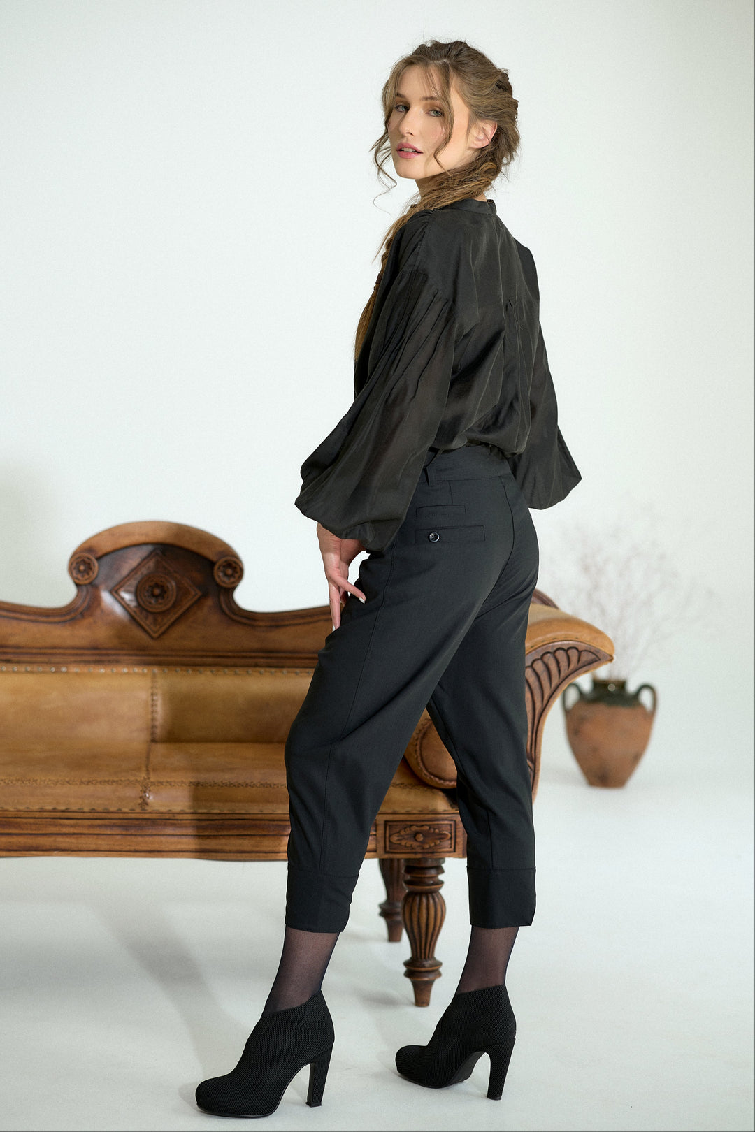 A woman posing in a black satin blouse and black cropped trousers, standing next to a vintage wooden bench in a studio setting.