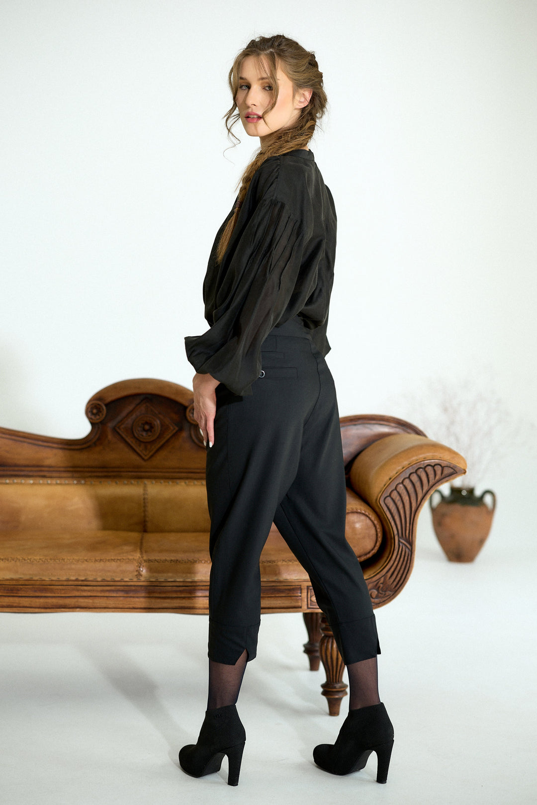 A woman posing in a black long-sleeve blouse and black cropped trousers, standing next to a brown leather chaise lounge in a studio setting.
