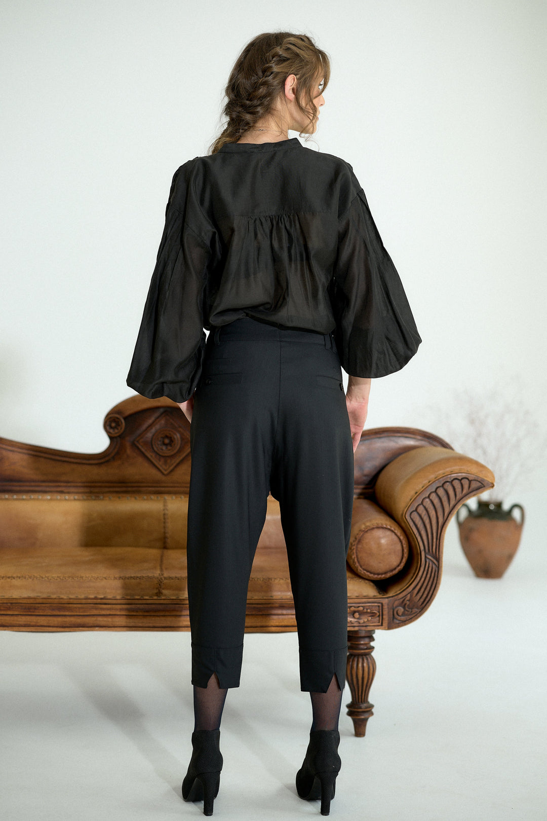 A woman stands with her back to the camera wearing a black sheer long-sleeve blouse with a gathered waist and black cropped trousers, posed in front of a brown leather chaise lounge against a white studio background.