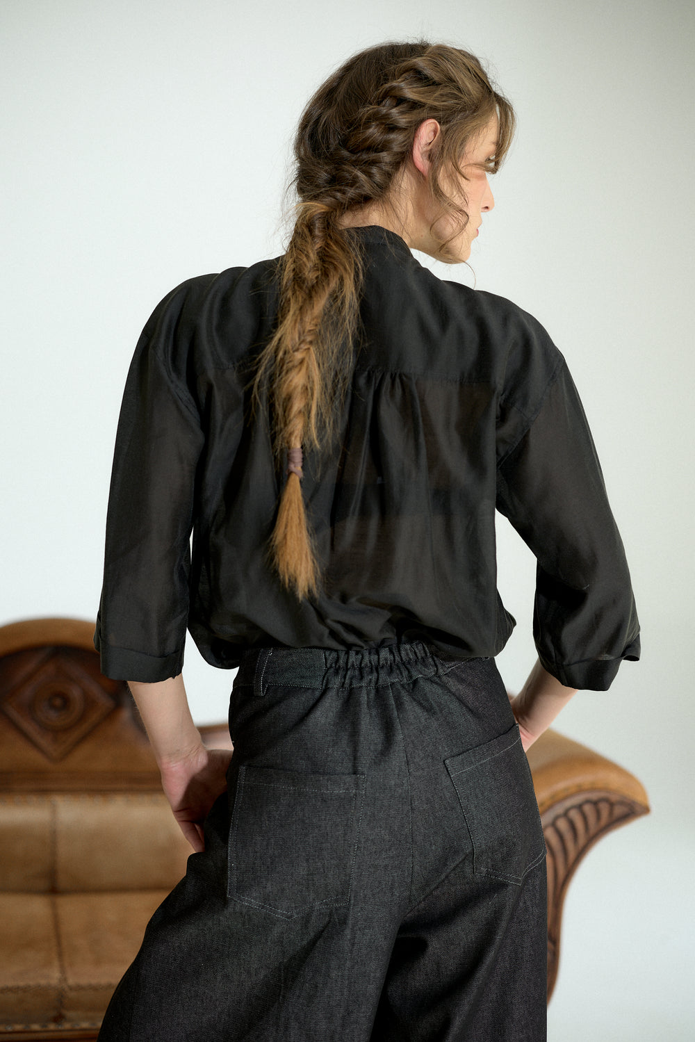 A woman shown from the back wearing a black long-sleeve blouse with a gathered neckline and dark charcoal wide-leg trousers, standing in front of a light gray wall with a brown leather chair partially visible.