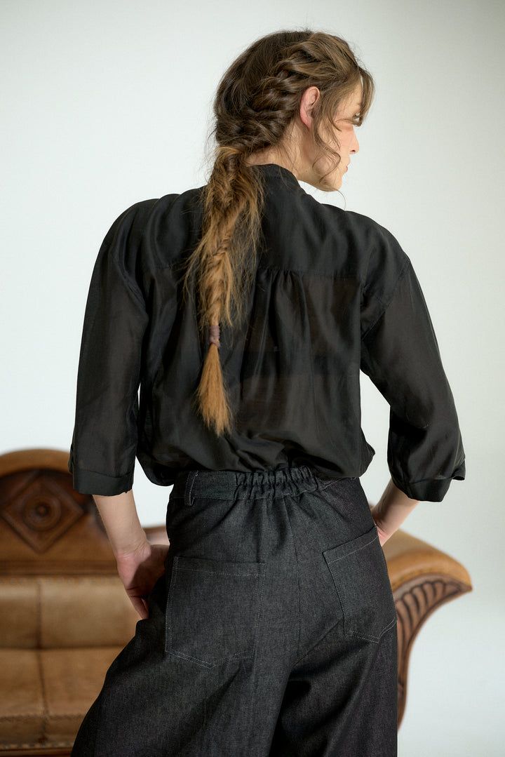 A woman shown from the back wearing a black long-sleeve blouse with a gathered neckline and dark charcoal wide-leg trousers, standing in front of a light gray wall with a brown leather chair partially visible.